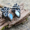 Bud shaped labradorite stones with tangled tendril tails. oxidised silver stud earrings with a dreamy elfin quality sits on a weathered piece of wood, the labradorite stones glinting blue in the sunlight. Shown with the buds facing down and the tendrils up.