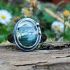 Oxidised silver ring with a lrage oval moss agate stone surrounded by a seaweed tendril. Close up showing the green inclusions in the clear agate stone and how it looks like a watercolour landscape. Sitting on a mossy log with daisys in the background.