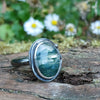 Oxidised silver ring with a large oval moss agate stone surrounded by a seaweed tendril. Side view showing the chunky D shape band. Sitting on a mossy log with daisys in the background.