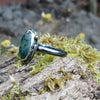 Oxidised silver ring with a large oval moss agate stone surrounded by a seaweed tendril. Side view showing the chunky D shape band. Sitting on a mossy log with daisys in the background.