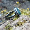 Oxidised silver ring with a large oval moss agate stone surrounded by a seaweed tendril. Side view showing the chunky D shape band. Sitting on a mossy log with daisys in the background.