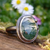 Oxidised silver ring with a lrage oval moss agate stone surrounded by a seaweed tendril. Close up showing the green inclusions in the clear agate stone and how it looks like a watercolour landscape. Sitting on a mossy log with daisys in the background.