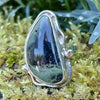 Sterling silver ring with a large green freeform oval Nebula Jasper stone surrounded with tendrils. The ring sits on a bed of moss.