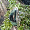 Sterling silver ring with a large green freeform oval Nebula Jasper stone surrounded with tendrils. The ring sits on a mossy log.