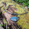 Oxidised black silver tendril pendant with blue flash labradorite bud winds around a bar and chain neckwire. Pictured on a mossy log. 
