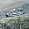 A sterling silver ring with a round, bezel-set watery blue Iolite gemstone, photographed close-up against a textured gray stone background.