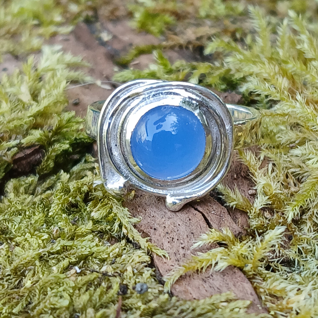 A silver ring with a round blue cabochon gemstone surrounded by silver tendrils photographed resting on a mossy log