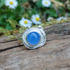 A silver ring with a round blue cabochon gemstone surrounded by silver tendrils photographed resting on a mossy log with daisys in the background
