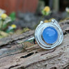 A silver ring with a round blue cabochon gemstone surrounded by silver tendrils photographed resting on a mossy log with blurred daisys in the background