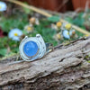 A silver ring with a round blue cabochon gemstone surrounded by silver tendrils photographed resting on a mossy log with blurred daisys in the background