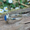 A silver ring with a round blue cabochon gemstone surrounded by silver tendrils photographed resting on a mossy log with blurred daisys in the background. side view showing slim silver band