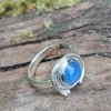 A silver ring with a round blue cabochon gemstone surrounded by silver tendrils photographed resting on a mossy log
