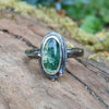 An oxidised silver ring with an oval green moss agate bezel set stone containing seaweed patterns, resting on a mossy log with a blurred garden background. The stone  is surrounded by seaweed tendrils which run down one side of the chunky D shaped band.