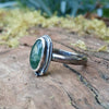 An oxidised silver ring with an oval green moss agate bezel set stone containing seaweed patterns, resting on a mossy log with a blurred garden background. The stone  is surrounded by seaweed tendrils which run down one side of the chunky D shaped band. Side view showing the curve of the stone