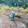 An oxidised silver ring with an oval green moss agate bezel set stone containing seaweed patterns, resting on a mossy log with a blurred garden background. The stone  is surrounded by seaweed tendrils which run down one side of the chunky D shaped band.