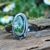 An oxidised silver ring with an oval green moss agate bezel set stone containing seaweed patterns, resting on a mossy log with a blurred garden background. The stone  is surrounded by seaweed tendrils which run down one side of the chunky D shaped band.