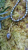 9ct yellow gold tendril pendant with a bezel set  bud shaped Labradorite stone on small black rice shaped pearls. The gold tendril loops around the small black freshwater pearl beads twice and curve back down to a point. resting on a mossy log in the sunshine.