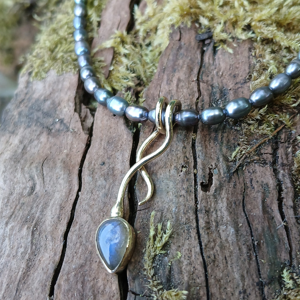 9ct yellow gold tendril pendant with a bezel set  bud shaped Labradorite stone on small black rice shaped pearls. The gold tendril loops around the small black freshwater pearl beads twice and curve back down to a point. resting on a mossy log with the stone flashing blue in the sunshine.
