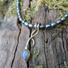 9ct yellow gold tendril pendant with a bezel set  bud shaped Labradorite stone on small black rice shaped pearls. The gold tendril loops around the small black freshwater pearl beads twice and curve back down to a point. resting on a mossy log with the stone flashing blue in the sunshine.