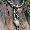 9ct yellow gold tendril pendant with a bezel set  bud shaped Labradorite stone on small black rice shaped pearls. The gold tendril loops around the small black freshwater pearl beads twice and curve back down to a point. resting on a mossy log with the stone flashing blue in the sunshine.