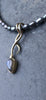 9ct yellow gold tendril pendant with a bezel set  bud shaped Labradorite stone on small black rice shaped pearls. The gold tendril loops around the small black freshwater pearl beads twice and curve back down to a point. taken against a slate background.