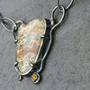 A close-up of a handmade pendant necklace featuring a natural ocean jasper stone with brown and cream patterns, set in a dark oxidised silver frame with a small yellow citrine gemstone, resting on a dark gray slate surface.