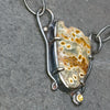 A close-up of a handmade pendant necklace featuring a natural ocean jasper stone with brown and cream patterns, set in a dark oxidised silver frame with a small yellow citrine gemstone, resting on a dark gray slate surface.