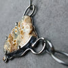 A close-up of a handmade pendant necklace featuring a natural ocean jasper stone with brown and cream patterns, set in a dark oxidised silver frame with a small yellow citrine gemstone, resting on a dark gray slate surface.