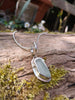 Long slim blue green seaglass pendant on black freshwater pearls draped over a mossy woodland log.