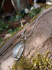 Long slim blue green seaglass pendant on smooth silver snake  chain draped over a mossy woodland log.