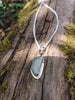 Long slim blue green seaglass pendant on chunky spiga chain lying on a mossy woodland log.