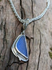 Triangle-ish  deep blue seaglass pendant with tendril detail on thick spiga chain on a mossy wooodland log. Close up.