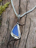 Triangle-ish deep blue seaglass pendant with tendril detail on thick spiga chain on a mossy wooodland log. Close up.