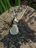 Round-ish olive seaglass pendant with tendril detail on smooth slim snake chain chain draped on a mossy woodland log.