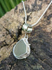 Round-ish olive seaglass pendant with tendril detail on smooth slim snake chain chain draped on a mossy woodland log. Close up.
