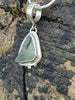 Small triangle-ish mid green seaglass pendant with tendril detail on smooth slim snake chain draped on a mossy woodland log. Close up.