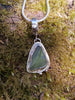Small triangle-ish mid green seaglass pendant with tendril detail on smooth slim snake chain draped on a mossy rock. Close up. 