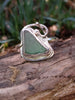 Front view of sterling silver ring with  teal green seaglass triangle  surrounded by  seaweed tendril detail which wanders down onto the shoulder of the D shaped band, sitting on a mossy log. 
