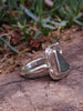 Side view of sterling silver ring with  teal green sea glass triangle  surrounded by  seaweed tendril detail which wanders down onto the shoulder of the D shaped band, sitting on a mossy log. 