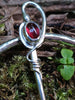 Traditional Silver brooch, penannular pin, ring pin with red and pink stones sits on a mossy log. Close up of the hilt of the pin set with a red garnet berry and a tendril twist.