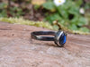 Black sterling silver ring with a teardrop shaped stone with a blue flash, sits on a mossy log in the sunshine.