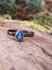 Black sterling silver ring with a teardrop shaped stone with a blue flash, sits on a mossy log in the sunshine.