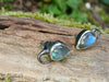 Bud shaped with tendril tails oxidised silver stud earrings with a dreamy elfin quality sits on a mossy log, the labradorite stones glinting blue in the sunlight.
