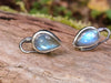 Bud shaped with tendril tails oxidised silver stud earrings with a dreamy elfin quality sits on a mossy log, the labradorite stones glinting blue in the sunlight.