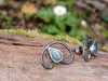 Bud shaped with tendril tails curling around, oxidised silver stud earrings with a dreamy elfin quality sits on a mossy log, the labradorite stones glinting blue in the sunlight.