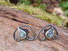Bud shaped with tendril tails curling around, oxidised silver stud earrings with a dreamy elfin quality sits on a mossy log, the labradorite stones glinting blue in the sunlight.