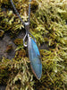 Large oval Labradorite and oxidised sterling silver pendant and long thick chain sitting on a mossy rock, the labradorite stone bud is clasped by black tendrils  and glints blue in the sunlight.