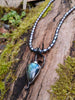 Labradorite bud and oxidised sterling silver pendant on a strand of lustrous black pearl beads sitting on a mossy log, glinting blue in the sunlight.