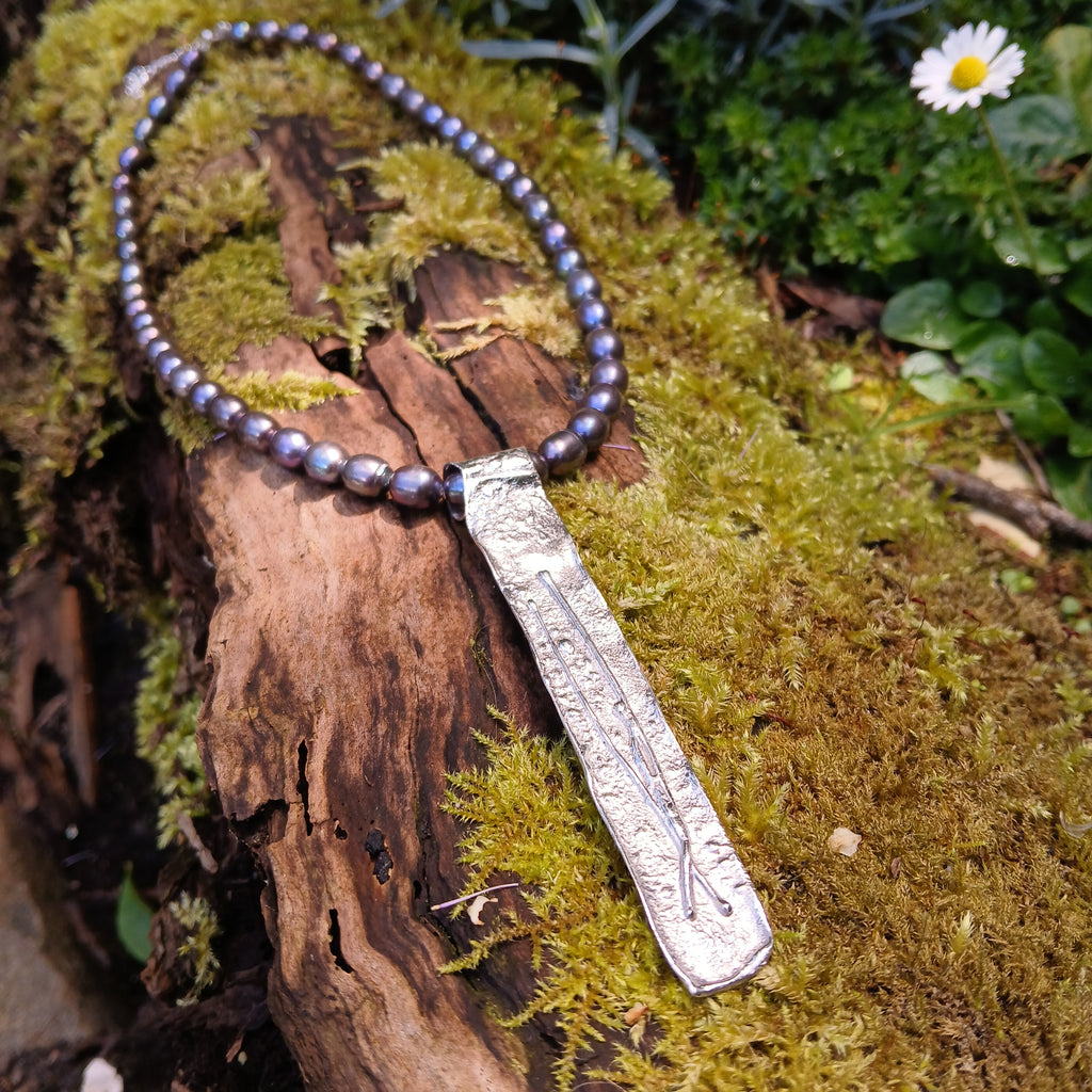Long Straight sterling silver melt textured pendant on black freshwater pearl beads. the necklace lies on a mossy woodland log.