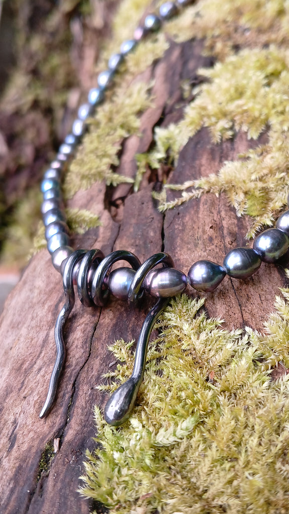 Oxidised silver spiral tendril pendant on black oval pearls, sitting on a mossy log in the sunshine